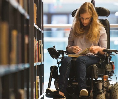 Opis slike: na slici je djevojka duge smeđe kose koja čita knjigu u biblioteci. Djevojka sjedi u motornim kolicima za osoba s invaliditetom, a pored nje se nalazi polica sa knjigama. Izvor slike je kampanja #PonosniNaSebe 